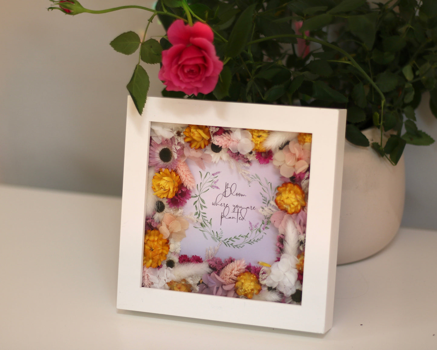 A white rectangular floral picture frame with white, pink and orange dried wild flowers and a sign "Bloom where you are planted"