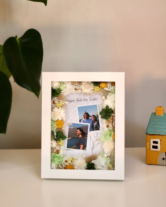 A sign "Happy birthday, Sister" in a white rectangular picture frame decorated with white and yellow flowers