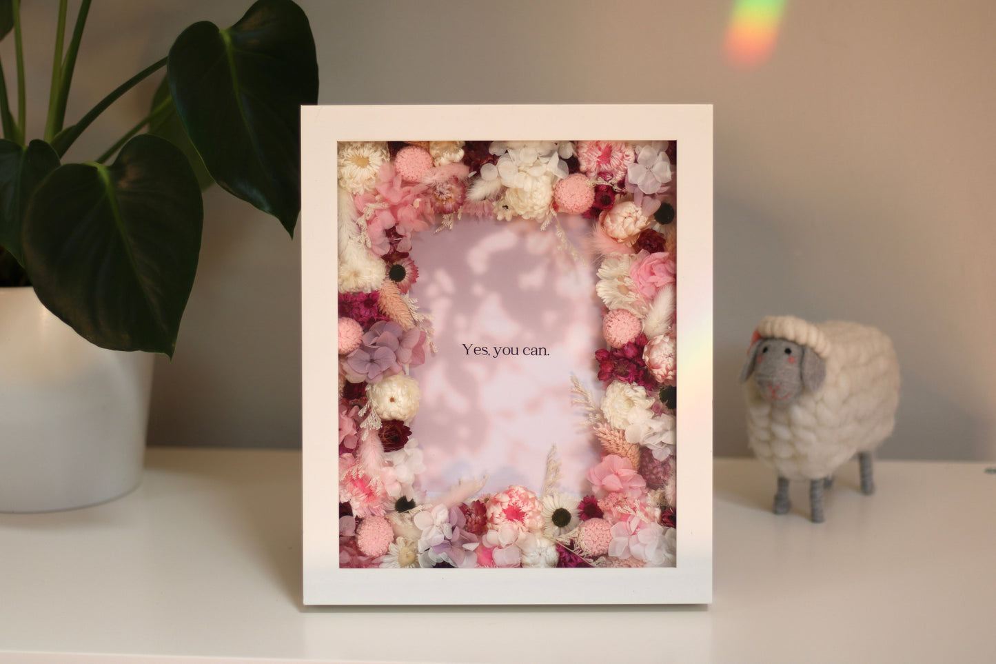 A white decorative picture frame with pink flowers and a motivational phrase Yes you can on a table near a little sheep behind