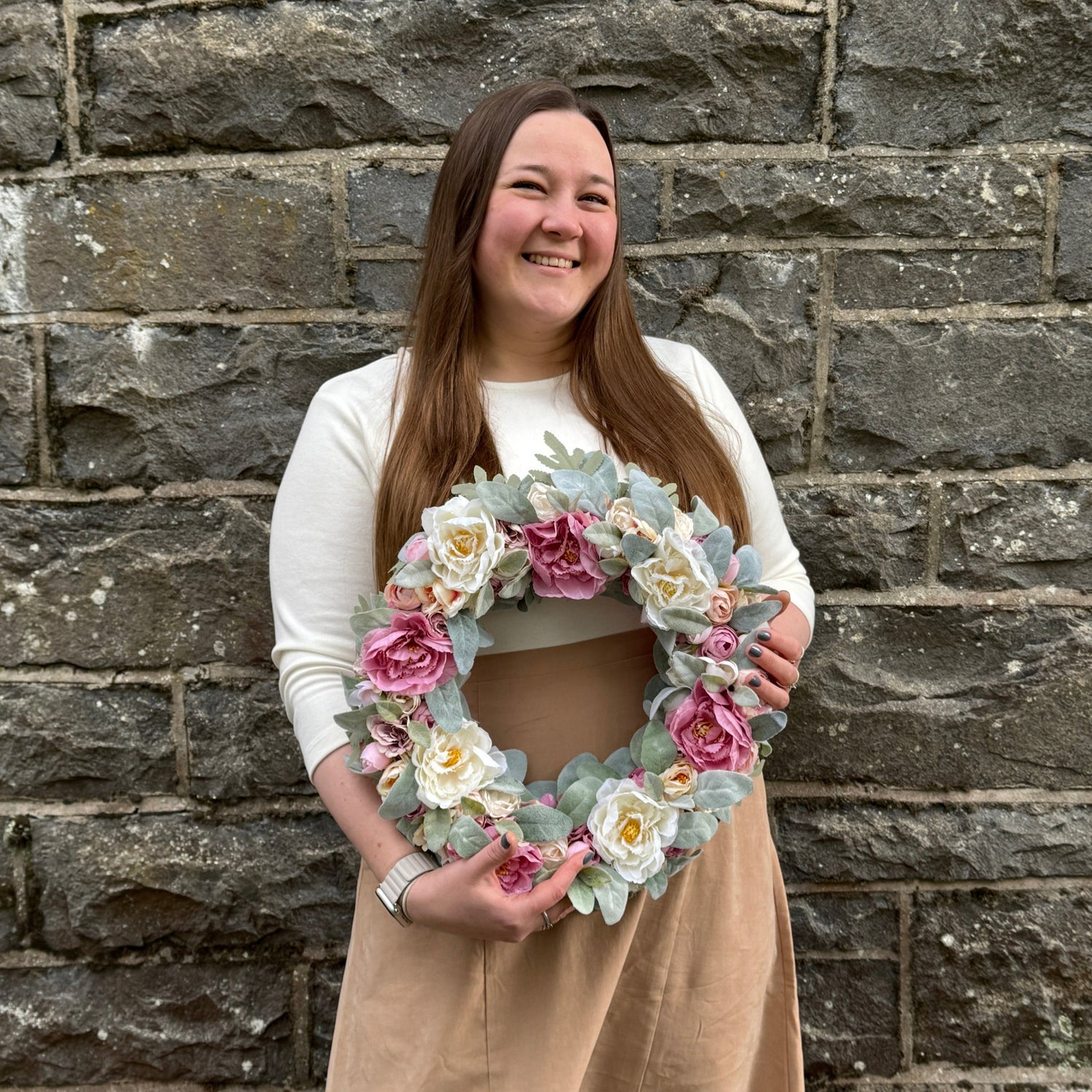Girl in front of a. grey wall holding a white and rose flower wreath