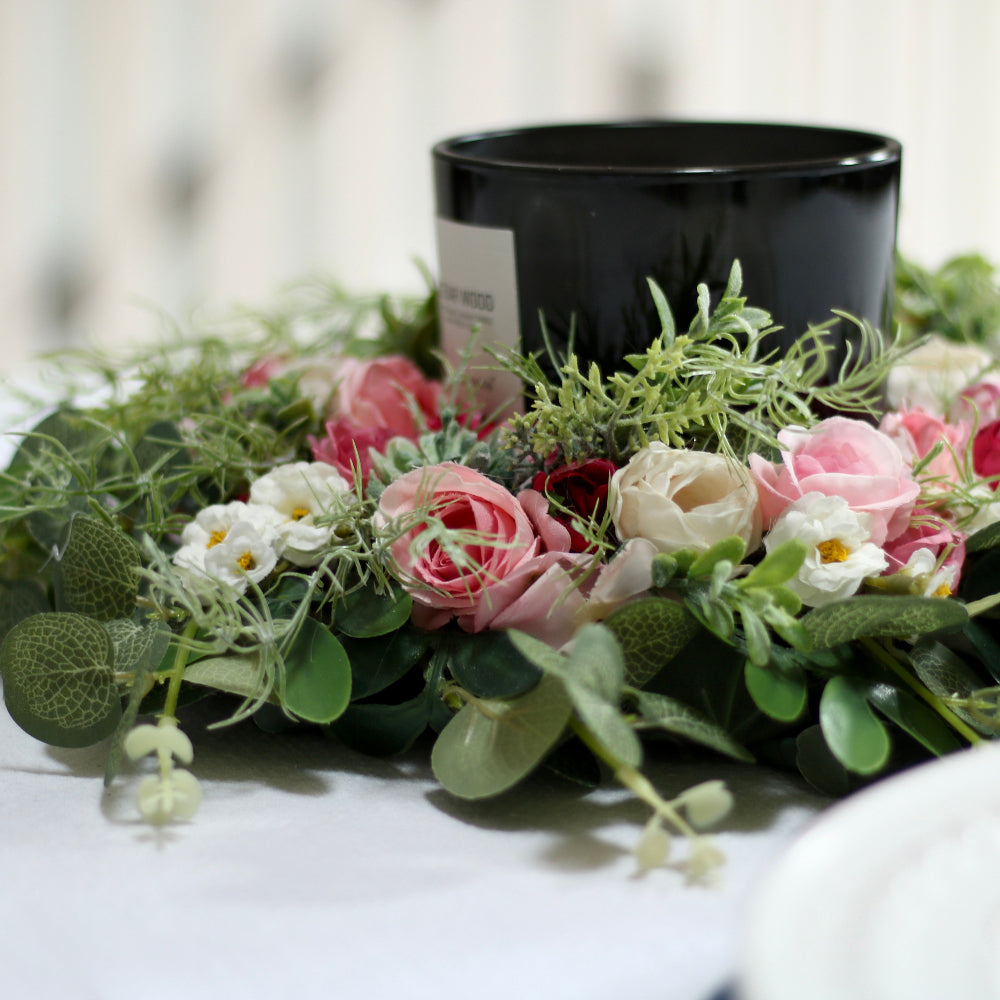 A wreath from pink flowers and green foliage with a black and white candle in the middle lies on a table next to a plate